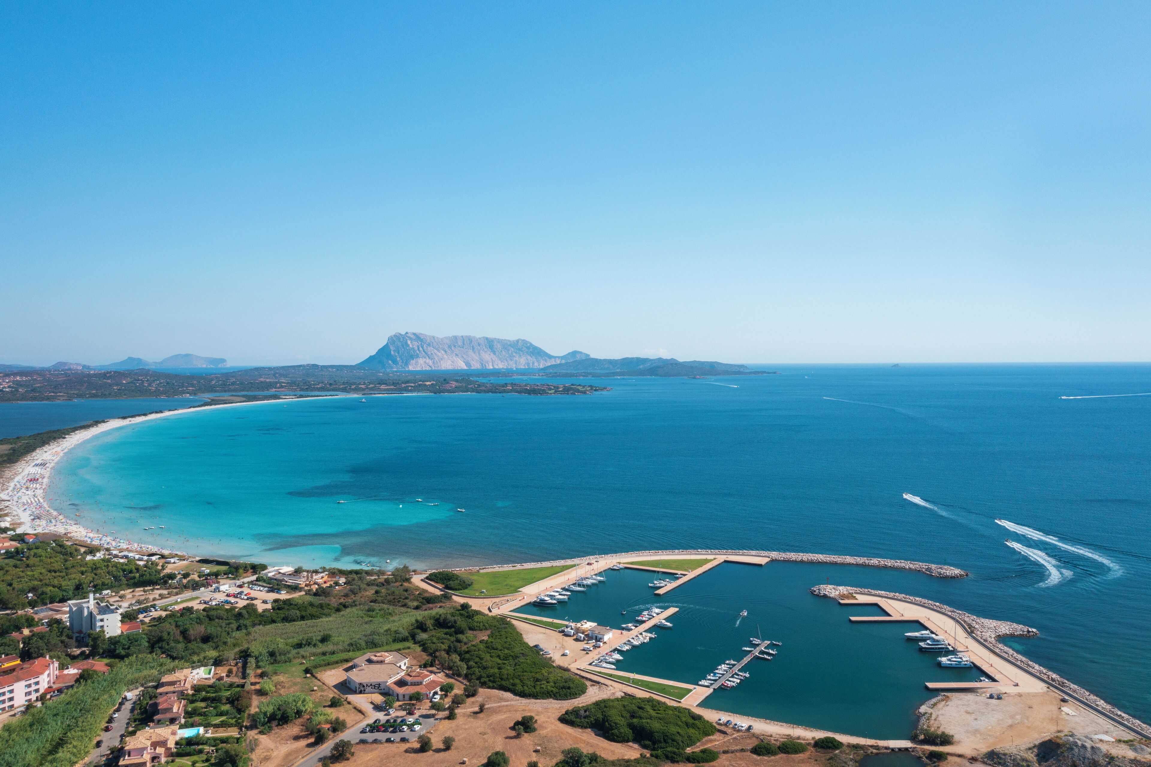 Un port de plaisance situé sur la côte, avec des bateaux amarrés, une plage de sable et une chaîne de montagnes en arrière-plan.