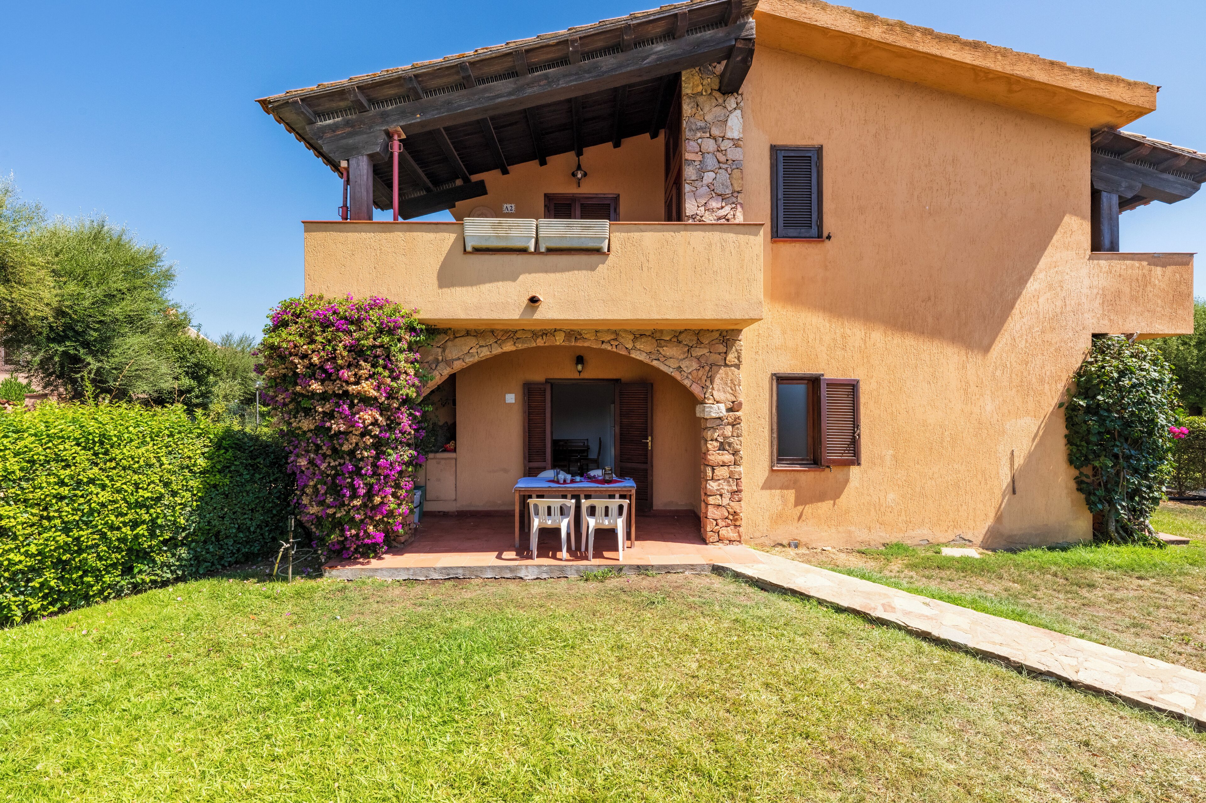 A two-story house with a stone archway entrance, a balcony, and a small patio with a table and chairs.