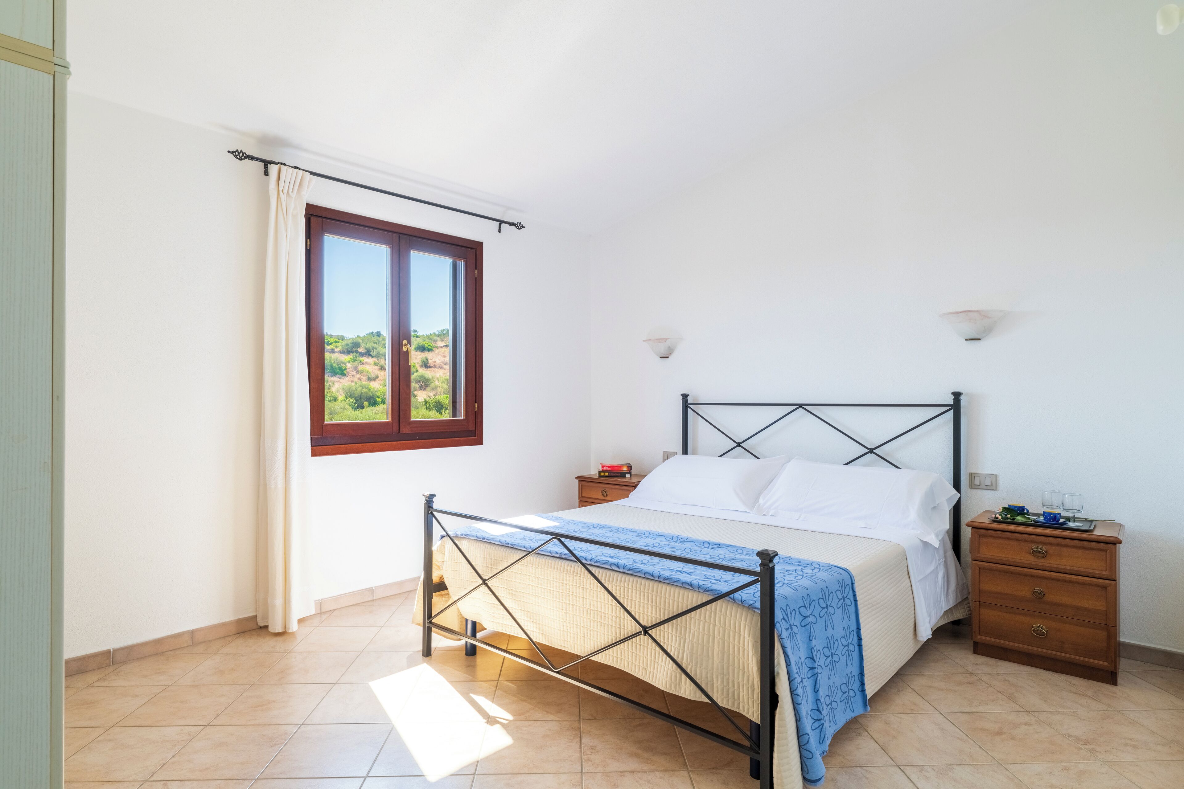 A bedroom with a bed, a nightstand, a window with curtains, and a view of greenery outside.
