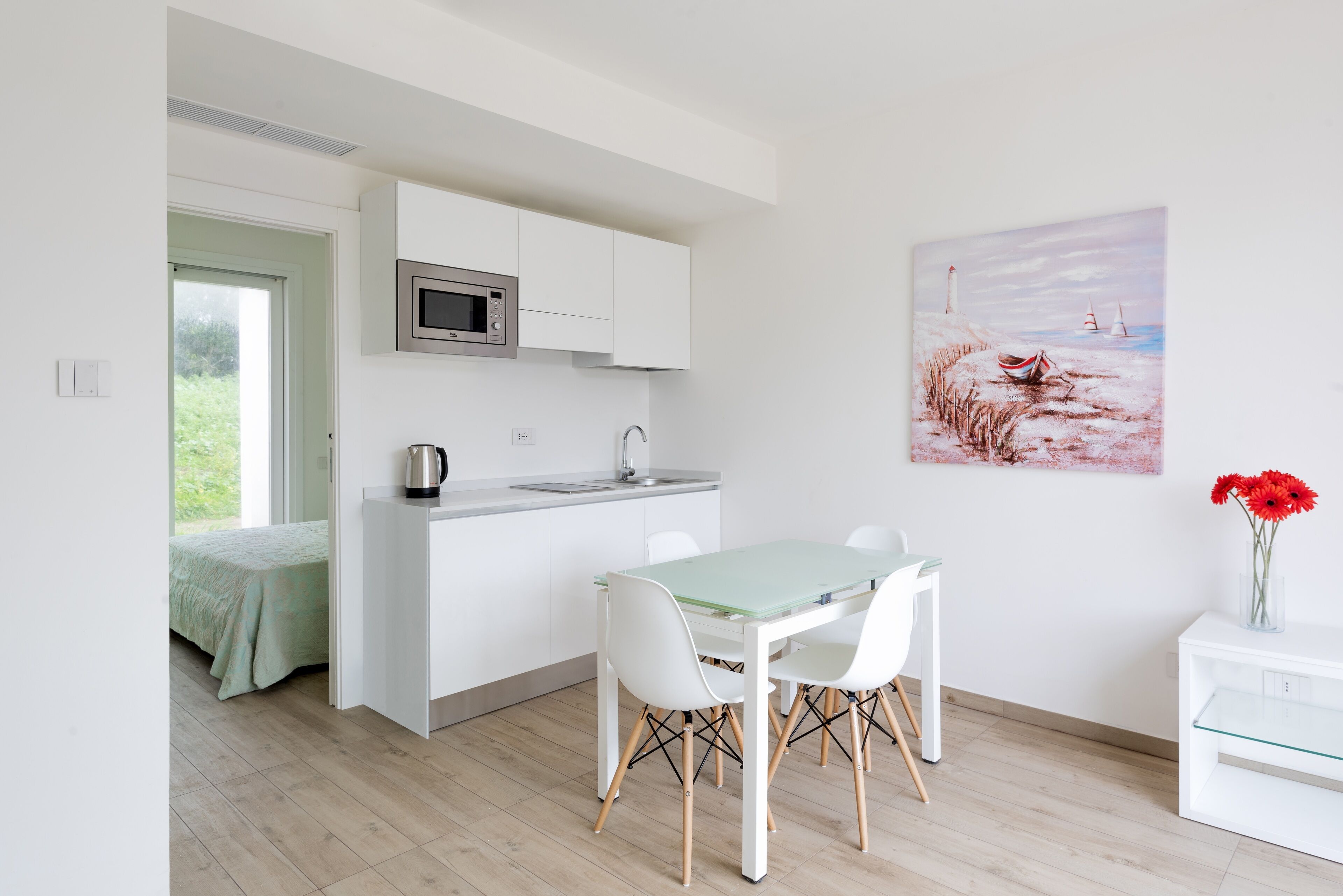 Une cuisine moderne avec des meubles blancs, un four à micro-ondes et un évier. On y trouve un coin repas avec une table en verre et des chaises blanches. Un tableau représentant un paysage côtier est accroché au mur.