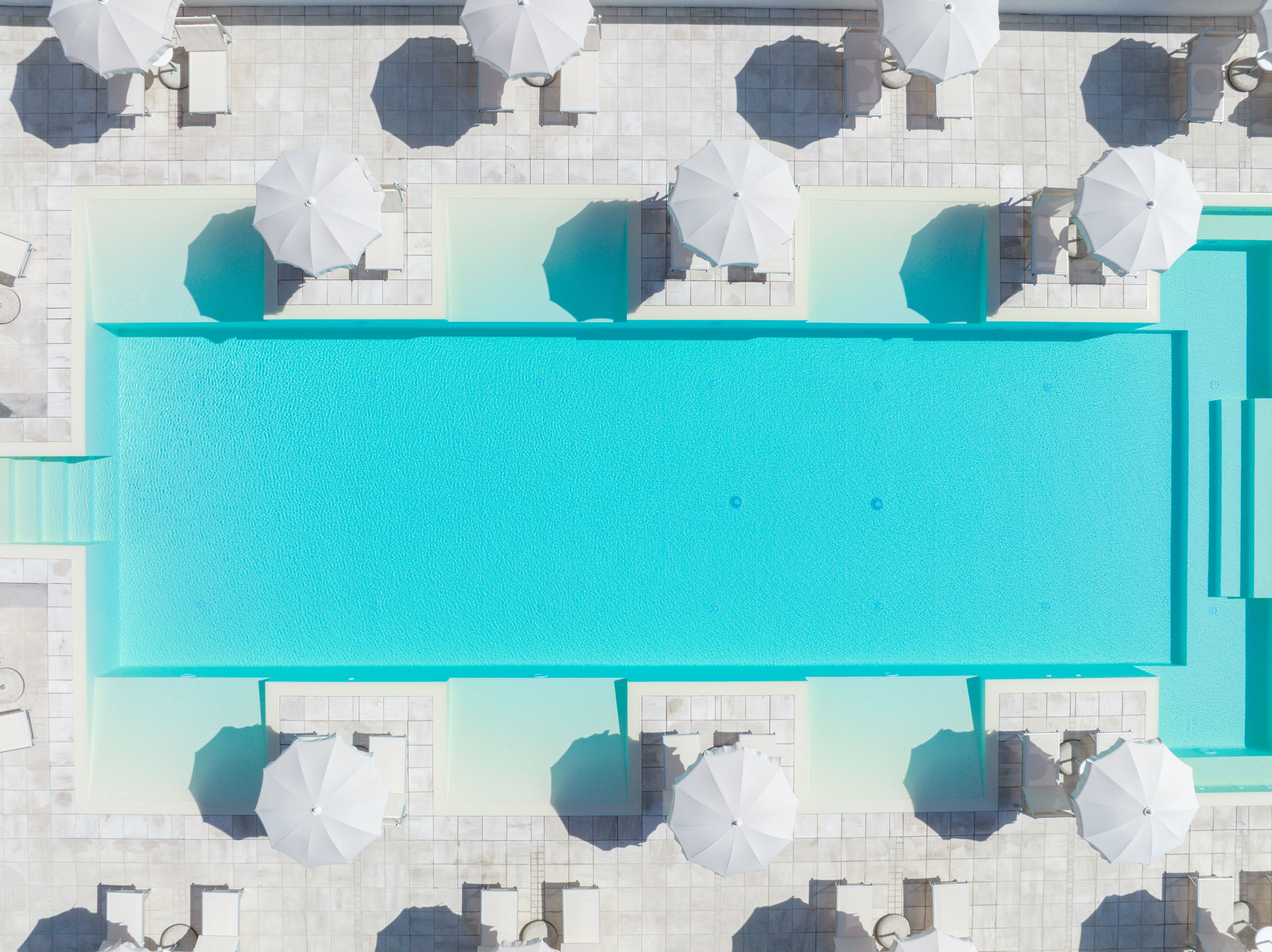 An aerial view of a rectangular swimming pool surrounded by white umbrellas and lounge chairs, indicating a resort setting.