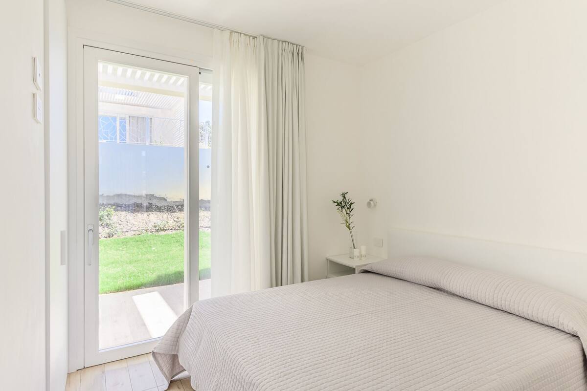 A bedroom with a bed, a nightstand, a vase with a plant, and a sliding glass door leading to a balcony.