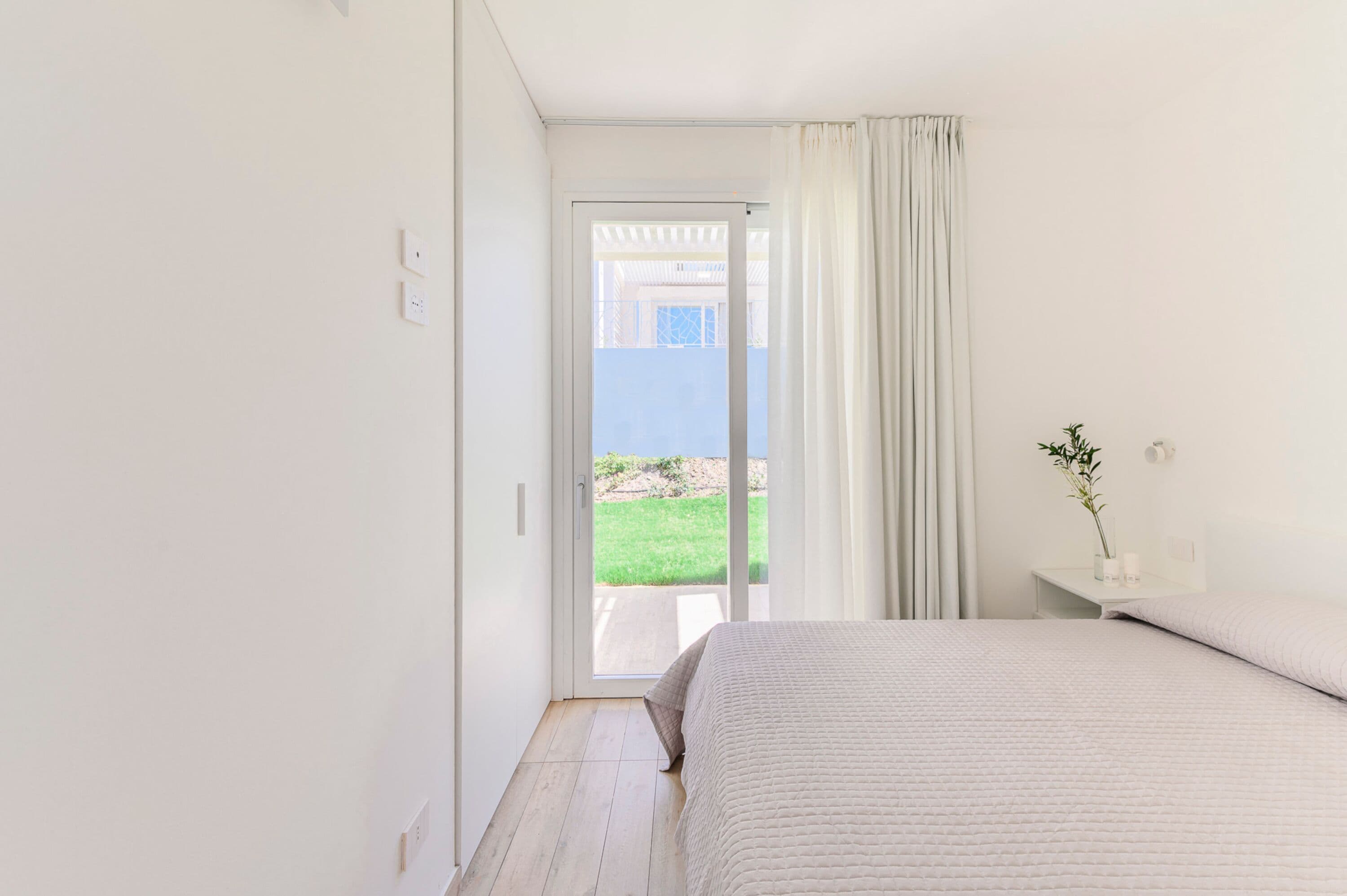 Ein Schlafzimmer mit einem Bett, einem Nachttisch mit einer Vase und Blick auf eine grüne Rasenfläche durch eine Schiebetür.