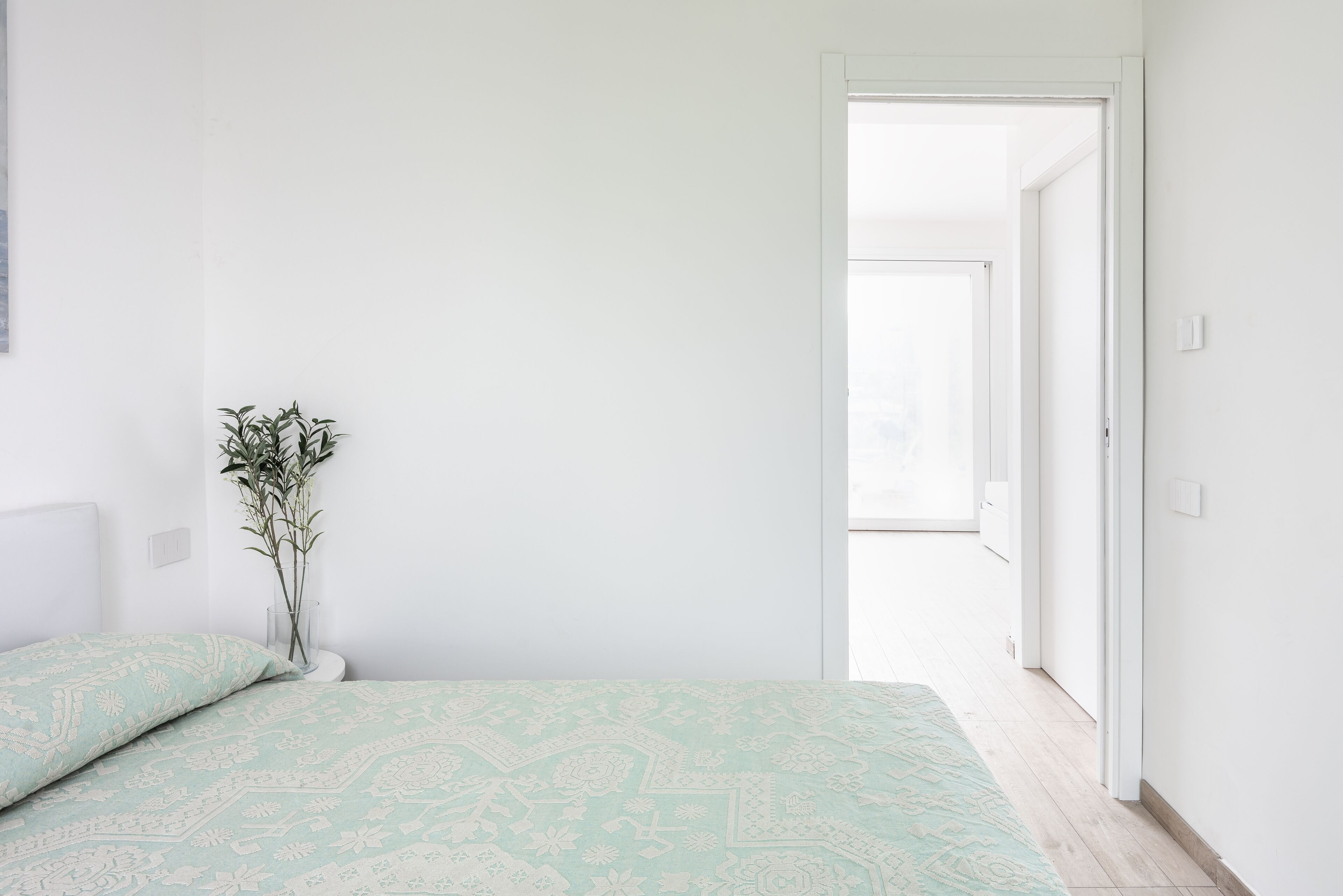 A bedroom with a bed, a vase with plants, and a door leading to another room.