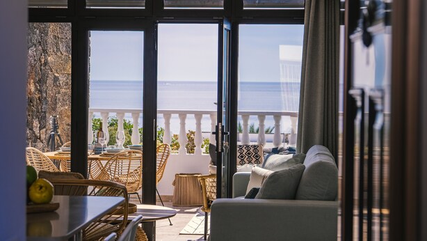 Een kamer met uitzicht op zee, een balkon met witte reling, rieten meubels en een bank met kussens.