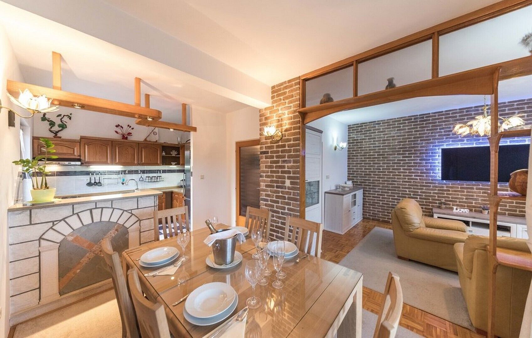 Una cocina moderna con armarios de madera y salpicadero de piedra, un comedor con pared de ladrillo y televisor, y un espacio de sala de estar acogedor con sofá y televisor.