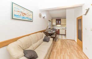 A living room with a beige sofa, a dining table with chairs, a kitchen in the background, and a framed painting on the wall.