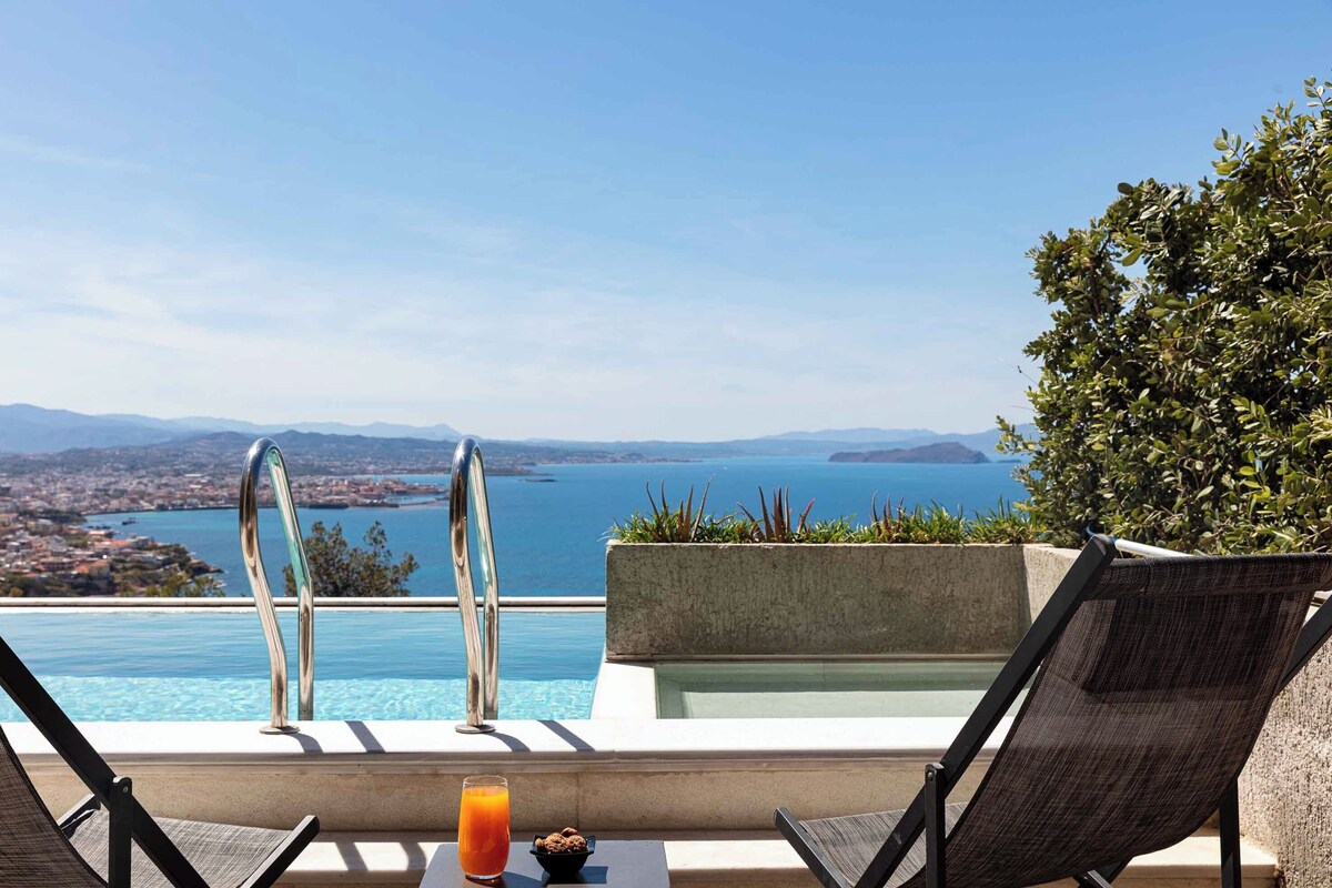 A poolside area with two lounge chairs, a table with a drink, and a view of the sea and distant landscape.