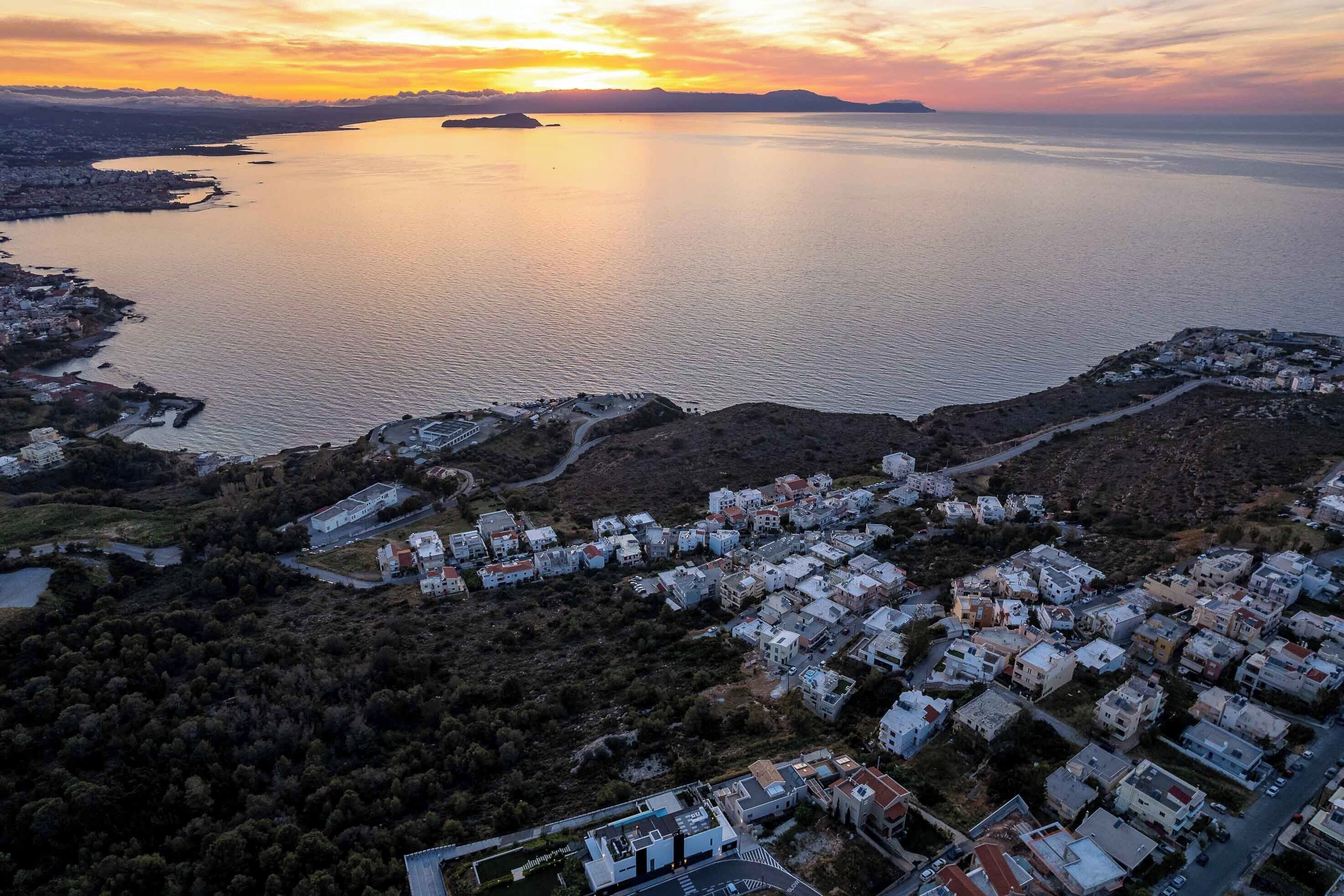 An aerial view of a coastal town with residential buildings, a marina, and a large body of water reflecting the sunset.
