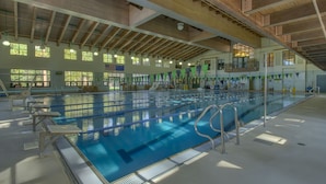 Un'ampia piscina coperta con diverse corsie, travi in legno a soffitto e finestre che lasciano entrare la luce naturale.