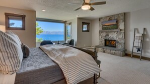 A bedroom with a large bed, a stone fireplace, a view of the sea, and a ceiling fan.
