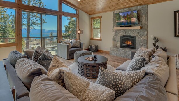 A spacious living room with a stone fireplace, large windows offering a view of the outdoors, and a cozy seating area. - California Sunshine - Lakeview - TLUXP (Carnelian Bay)