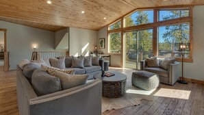 A spacious living room with a large gray sectional sofa, a round coffee table, and a patterned ottoman. The room features wooden flooring, a sloped ceiling with exposed beams, and large windows offering a view of trees and a building outside. - California Sunshine - Lakeview - TLUXP (Carnelian Bay)