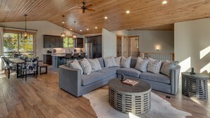A spacious living area with a large grey sectional sofa, a round coffee table, and a dining table with chairs. The room features wooden flooring, a high ceiling with a fan, and large windows allowing natural light. - California Sunshine - Lakeview - TLUXP (Carnelian Bay)