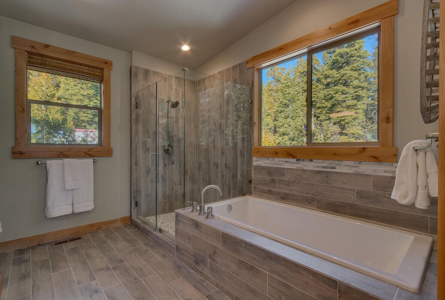 A modern bathroom with a glass-enclosed shower, a large bathtub, and two windows offering views of trees.