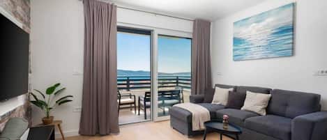 Un salon moderne avec un canapé gris, une petite table basse et un téléviseur sur le côté gauche. Un balcon avec vue sur la mer est visible à travers les portes vitrées.