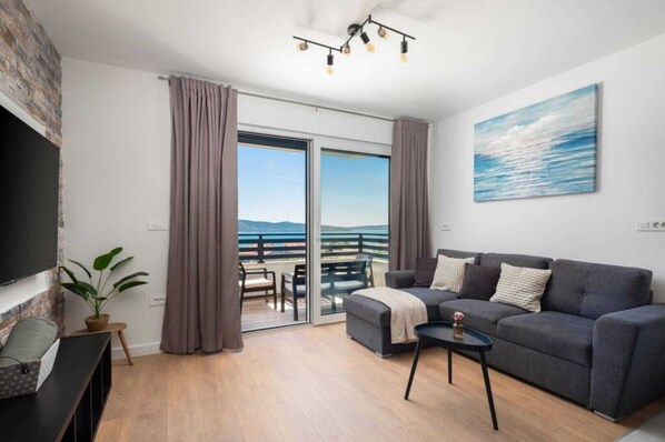 A modern living room with a grey sofa, a small coffee table, and a TV on the left. A balcony with a view of the sea is visible through the glass doors.