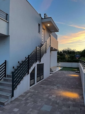 Un edificio moderno con una escalera que conduce a un segundo piso, techo plano y un área exterior pavimentada.