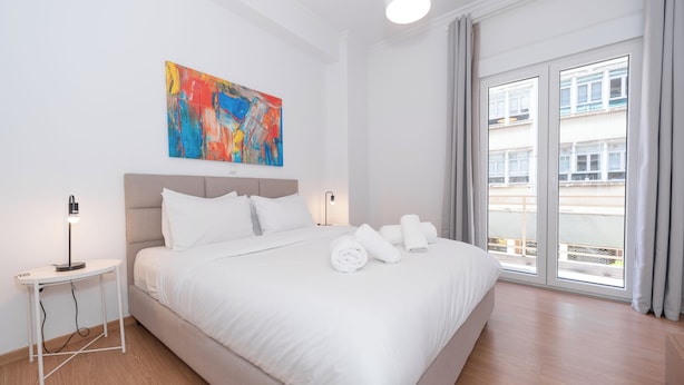 Une chambre d’hôtel moderne avec un grand lit, une table de chevet avec une lampe, une peinture abstraite aux couleurs vives accrochée au mur, et une vue sur la ville par la fenêtre.