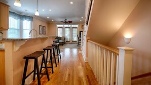 A kitchen with wooden stools, a dining area with a sofa, and a staircase with a railing. - Blue Bliss - 6 Bedroom Beach House -1 Block from Beach Access. Linens Included! (Carolina Beach)