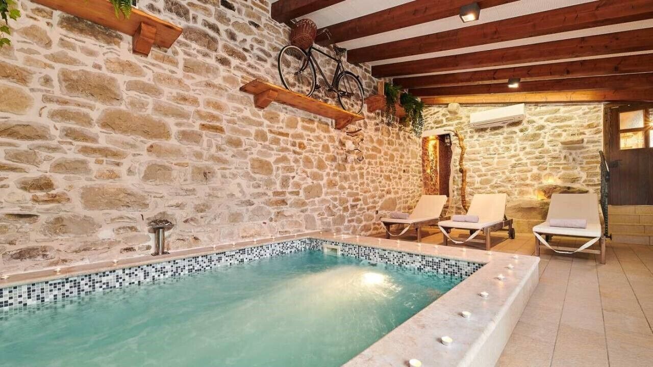 A stone-walled indoor pool with a tiled edge, wooden beams, and two lounge chairs with cushions.