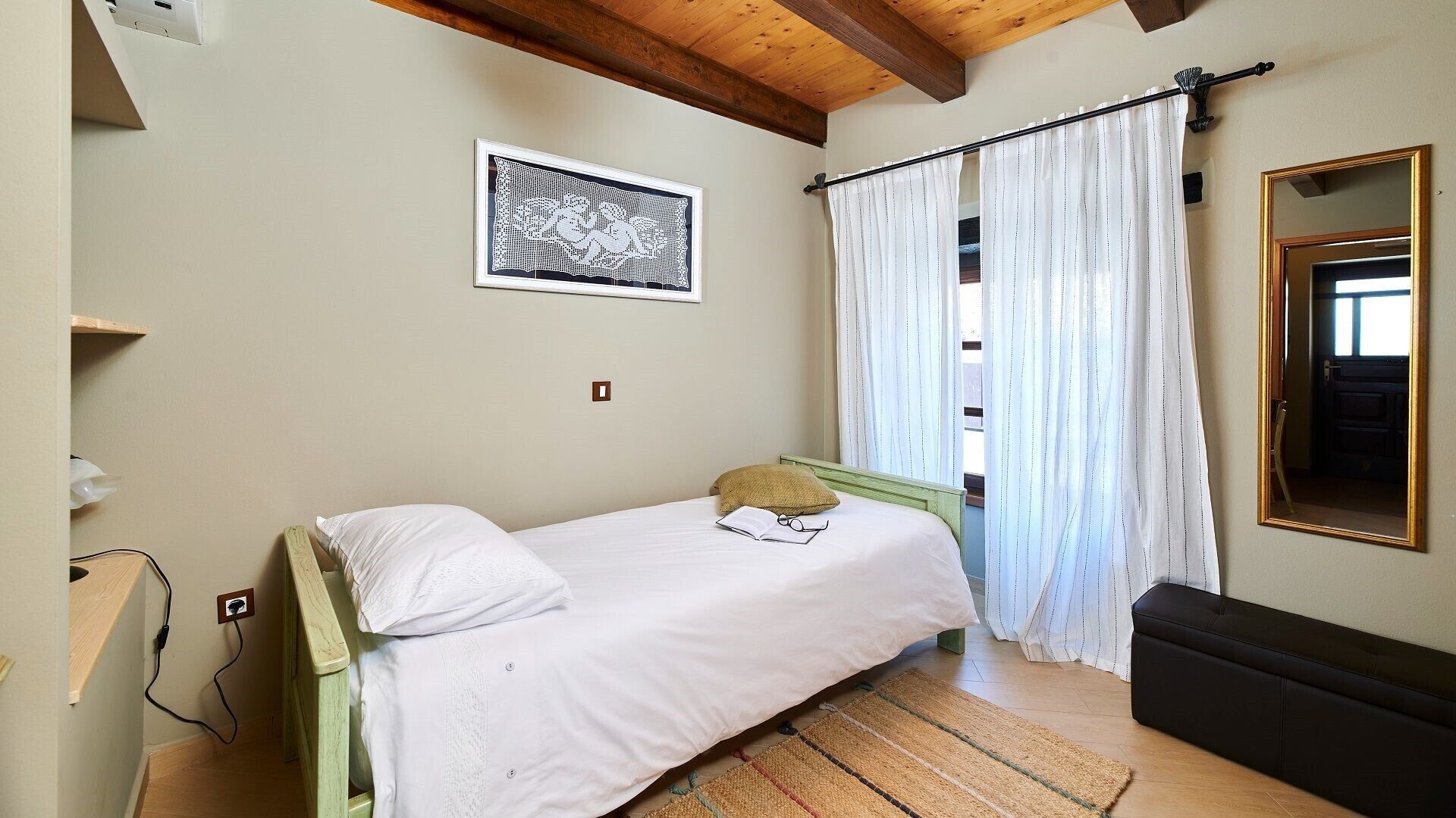 Uma cama de solteiro com colchão e almofadas brancos, uma cabeceira de madeira, um tapete tecido, uma pequena mesa de cabeceira com abajur, um quadro emoldurado na parede e uma janela com cortinas brancas.