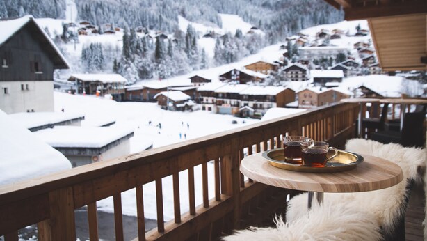 Un balcón de madera con una mesa y sillas, con vistas a un pueblo cubierto de nieve, casas y una montaña al fondo.
