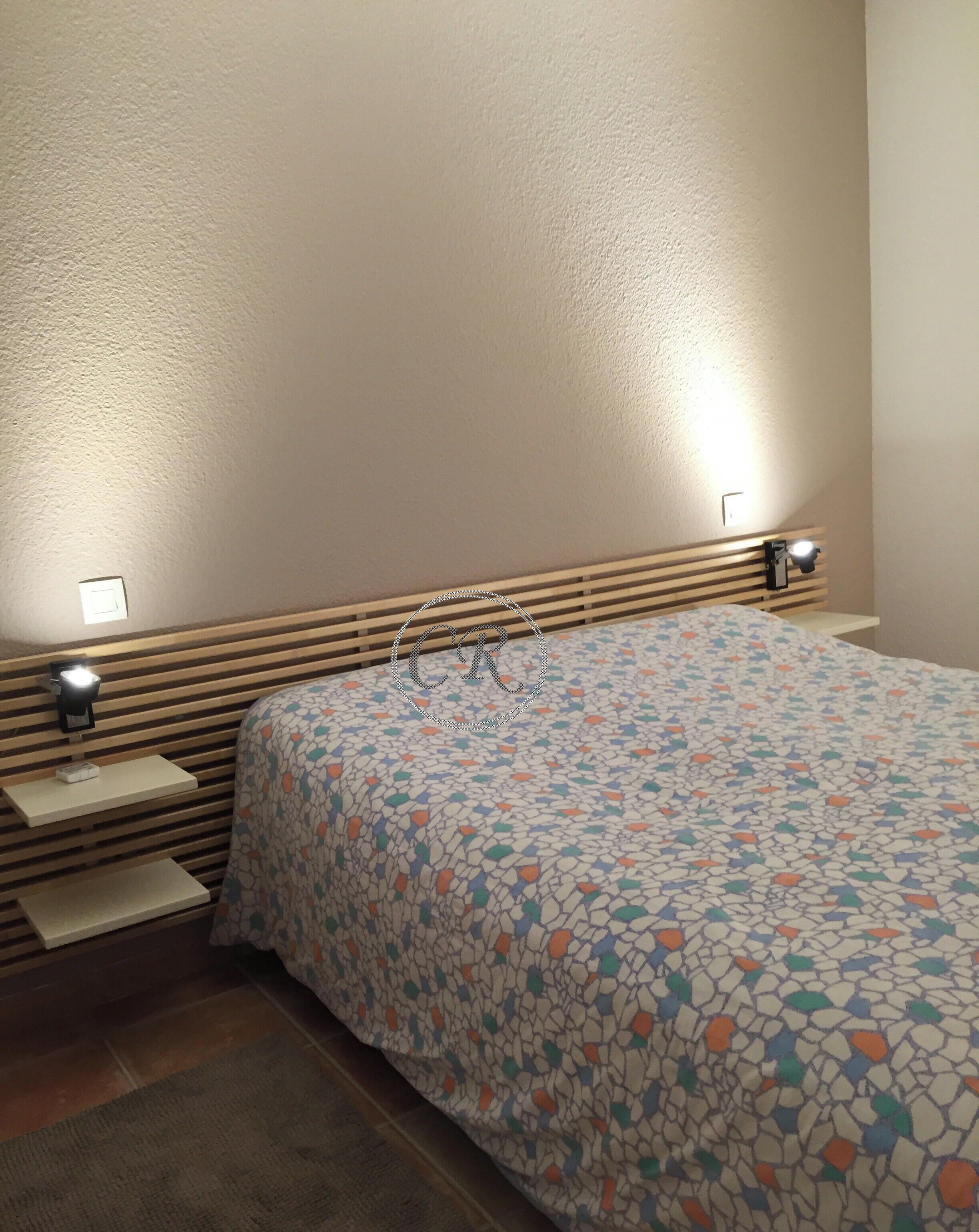 A bedroom with a bed, two bedside tables, a nightstand, a wall-mounted lamp, and a textured wall.