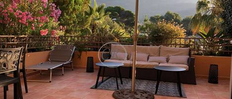 A patio with a sofa, chairs, and a table under a canopy, surrounded by lush greenery and a mountain view.