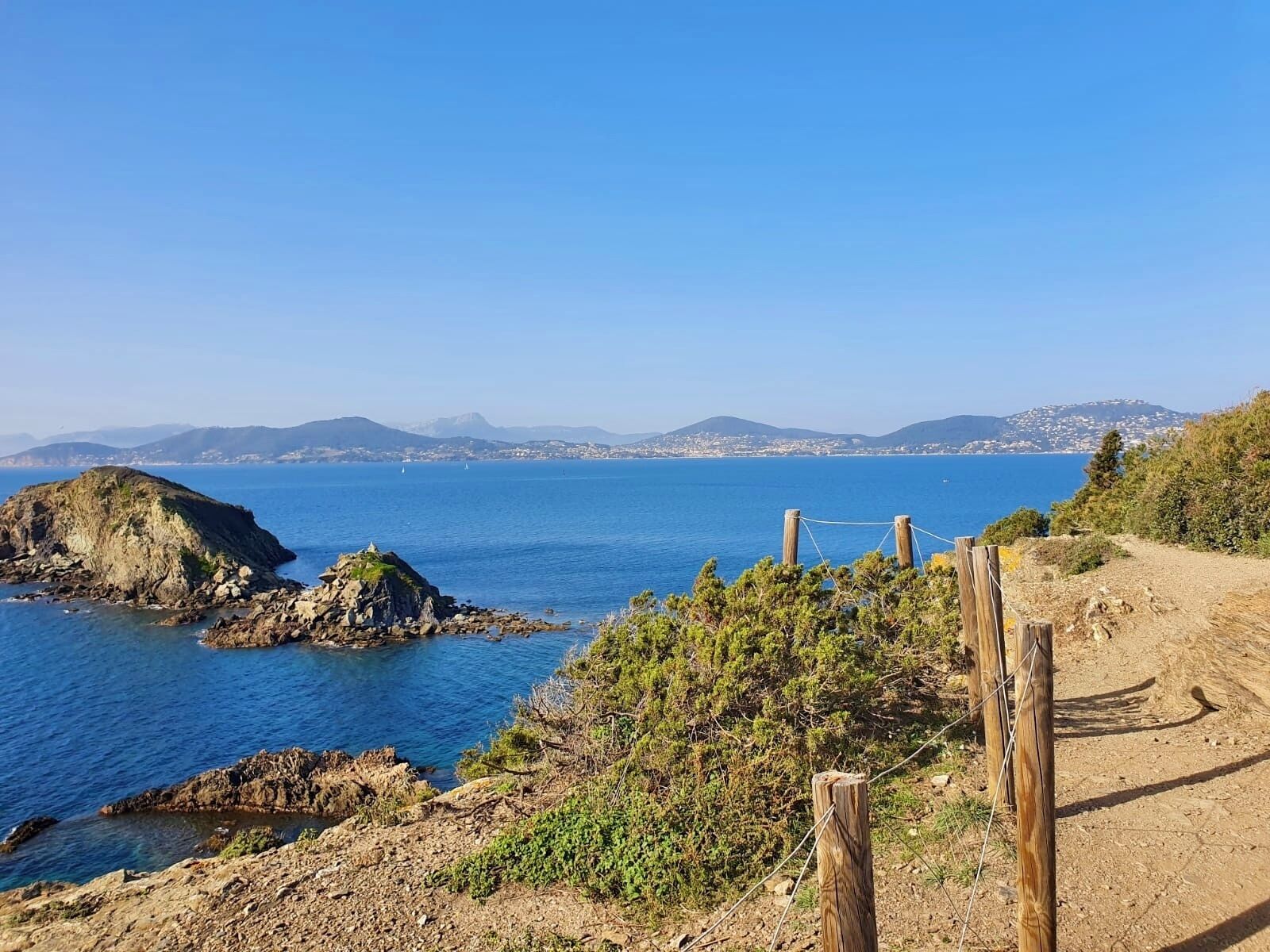 A coastal landscape with rocky terrain, a dirt path, and a clear view of the sea and distant hills.