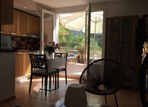 Una cocina con armarios de madera, un comedor con mesa y sillas, y vista a un jardín a través de un ventanal abierto.