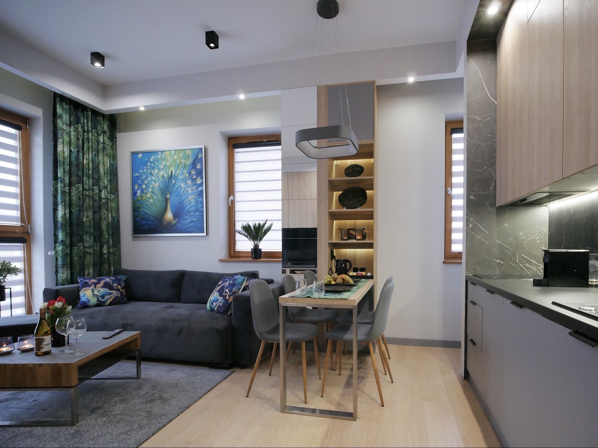 A modern living room with a dark grey sofa, a wooden coffee table, and a dining area with a table and chairs. There is a kitchen area with a stove and cabinets. A large painting of a peacock is on the wall, and a window with blinds is visible.
