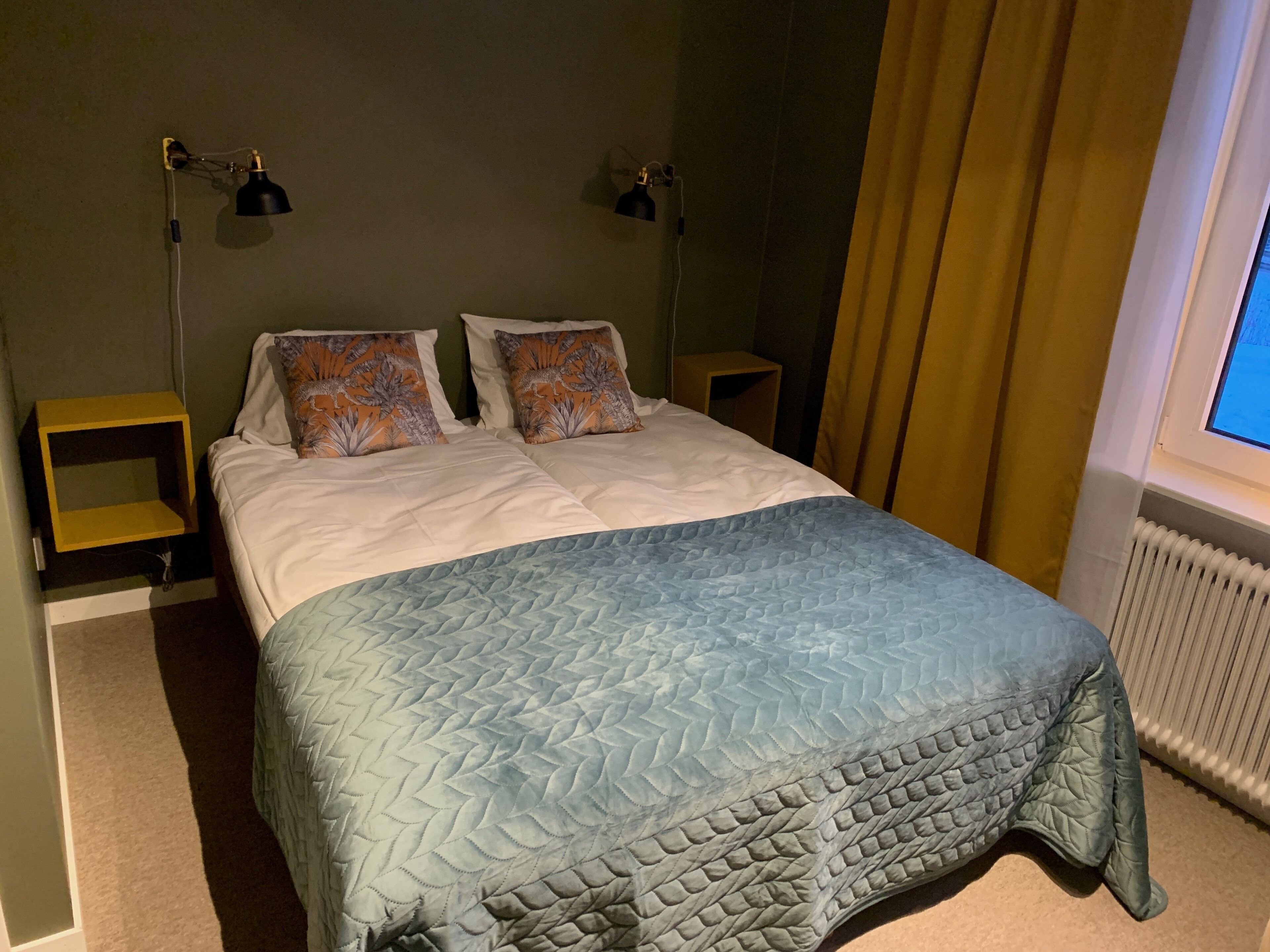 Een keurig opgemaakt bed met een gewatteerde spreidek, twee bedlampen en een raam met gordijnen.