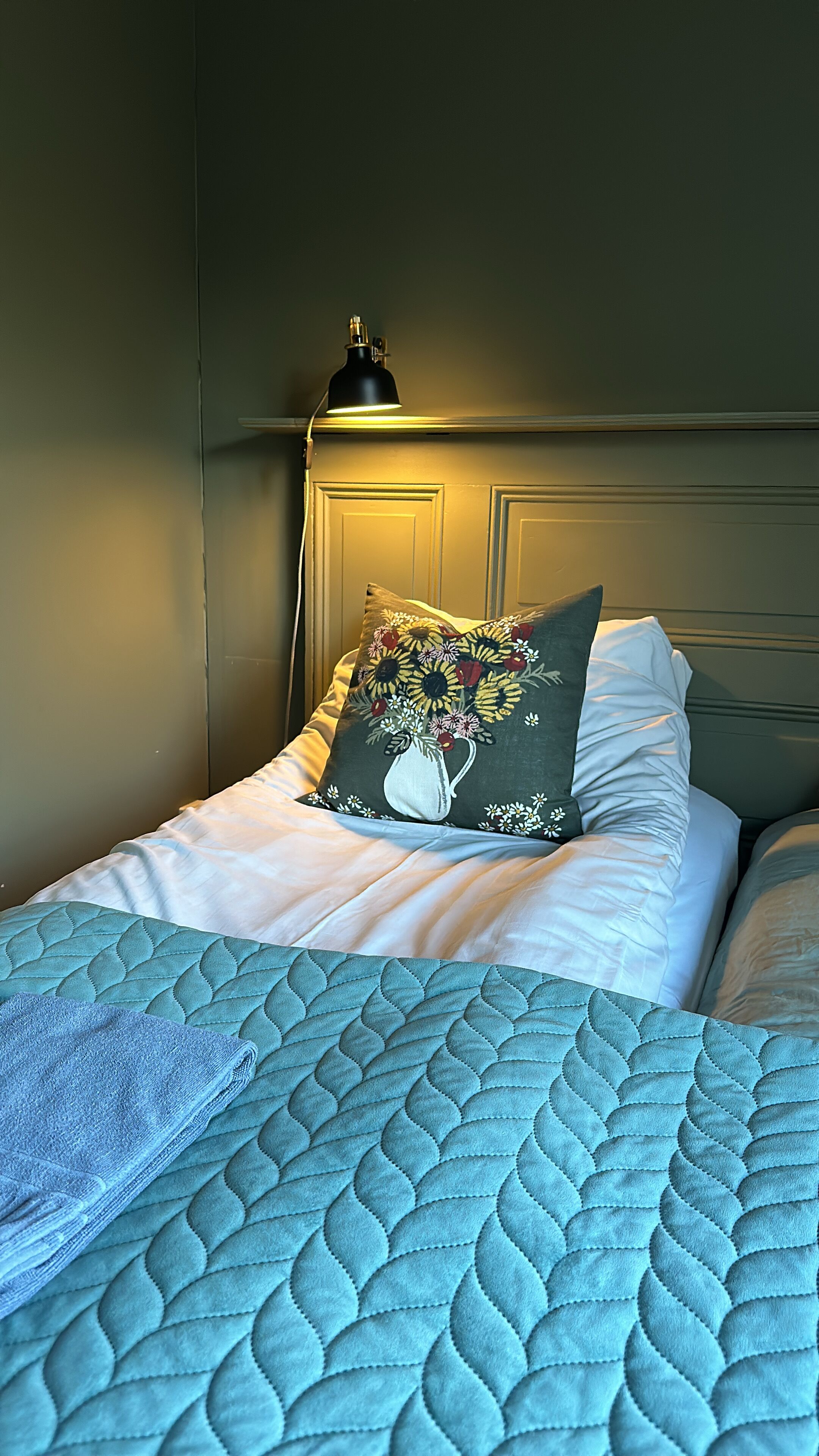 A neatly made bed with a blue quilt, a pillow with a floral design, and a bedside lamp.