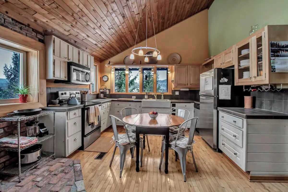 A kitchen with wooden ceiling, a dining table with chairs, and a view of trees through large windows.