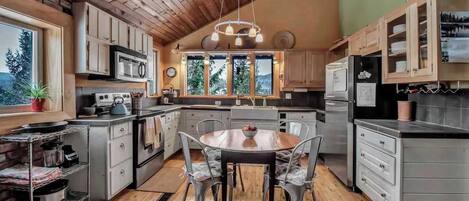 A kitchen with wooden ceiling, a dining table with chairs, and a view of trees through large windows.