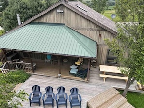 A wooden barn with a green metal roof, a covered porch with outdoor seating, and a wooden deck with chairs. - Rustic Cabin, Wood Fireplace & Large Deck (Great Valley)