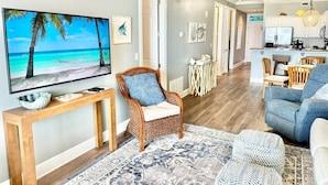 A living room with a TV, a wicker chair, a blue sofa, a dining area, and a kitchen in the background. - Premiere Complex. Beachfront Penthouse Unit W/ Amazing View, Extra Large Balcony (Gulf Shores)
