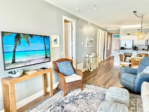 A living room with a TV, a wicker chair, a blue sofa, a dining area, and a kitchen in the background.