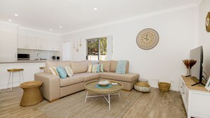 Ein modernes Wohnzimmer mit einem beigefarbenen Ecksofa, einem runden Kaffeetisch aus Rattan und einer Küche im Hintergrund.
