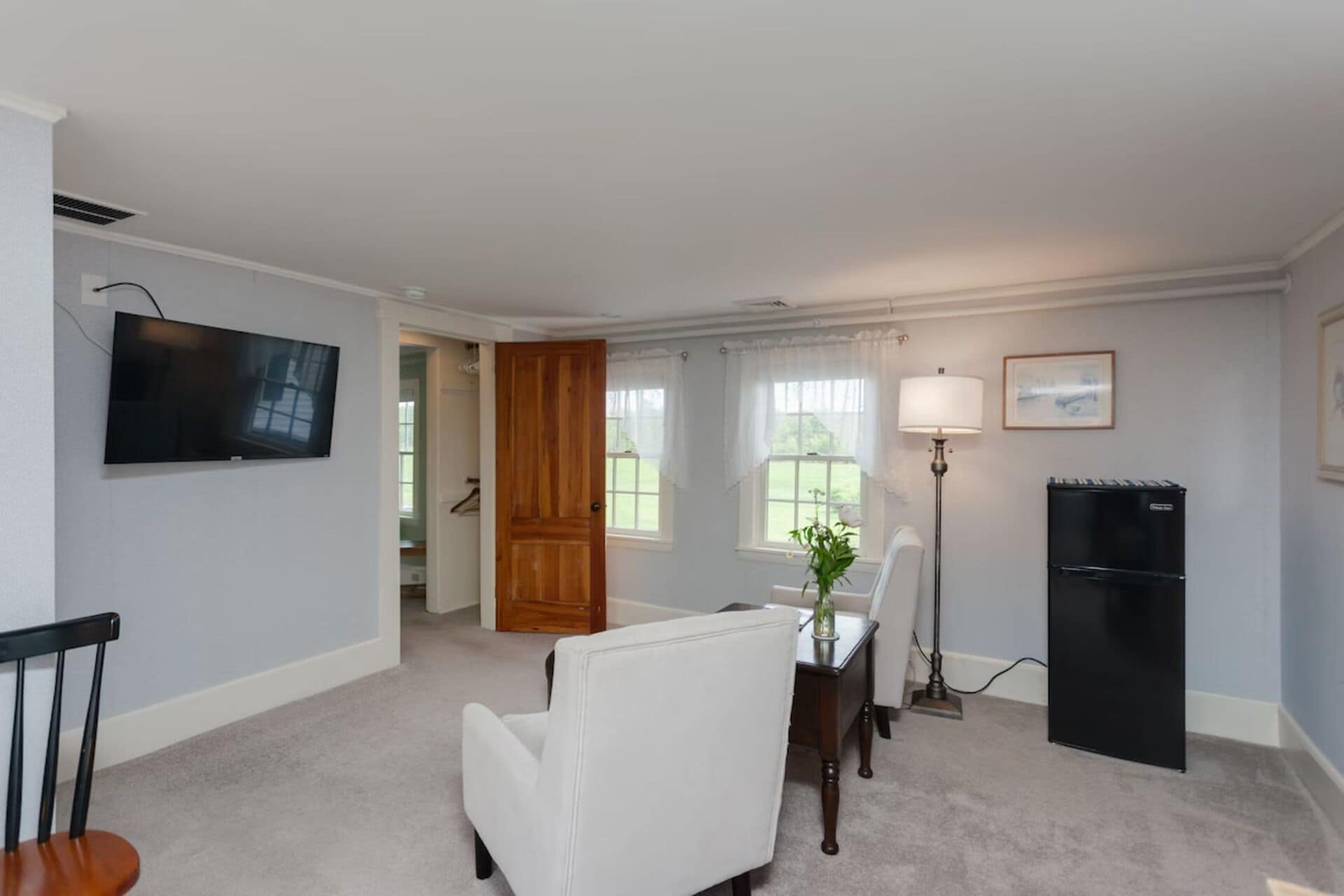 A room with a flat-screen TV, a dining table with chairs, a lamp, a refrigerator, and a door leading to another room.