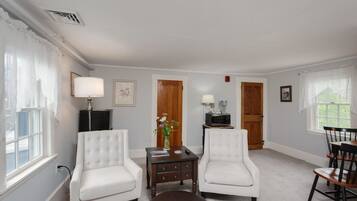A living room with two white armchairs, a small table with a vase, a television, and a wooden door.