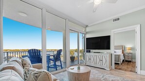 Un vaste espace de vie avec une grande porte-fenêtre coulissante donnant sur un balcon, un téléviseur à écran plat et un coin salon confortable avec une table basse ronde.