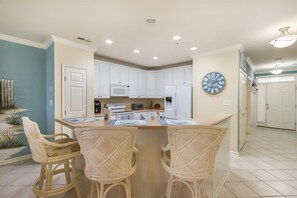 A kitchen with white cabinets, a microwave, a refrigerator, and a clock on the wall. There are wicker chairs around a small table.