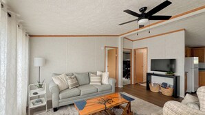 A living room with a sofa, coffee table, TV, and kitchen area. - Riverside Cottage w/ 70' Private Dock - 5 Minutes From Town! (Cheboygan)