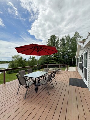 A wooden deck with a glass table and chairs, a red umbrella, and a view of a lake and trees.
