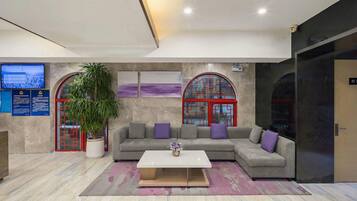 Un lobby moderno con un sofá modular, una mesa de centro, una planta grande en maceta y una pantalla de televisión.