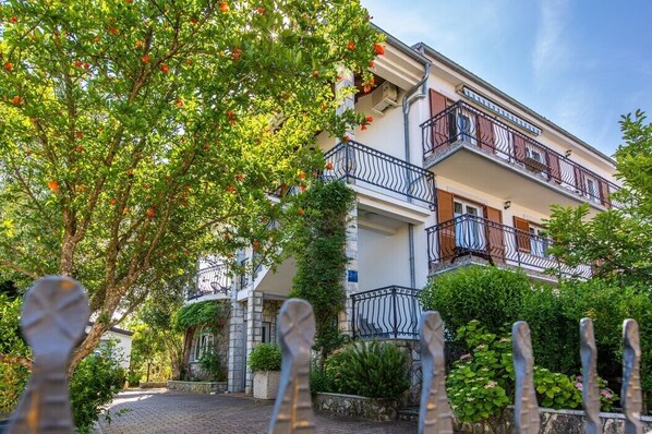 A multi-story building with balconies, surrounded by lush greenery and a cobblestone path.
