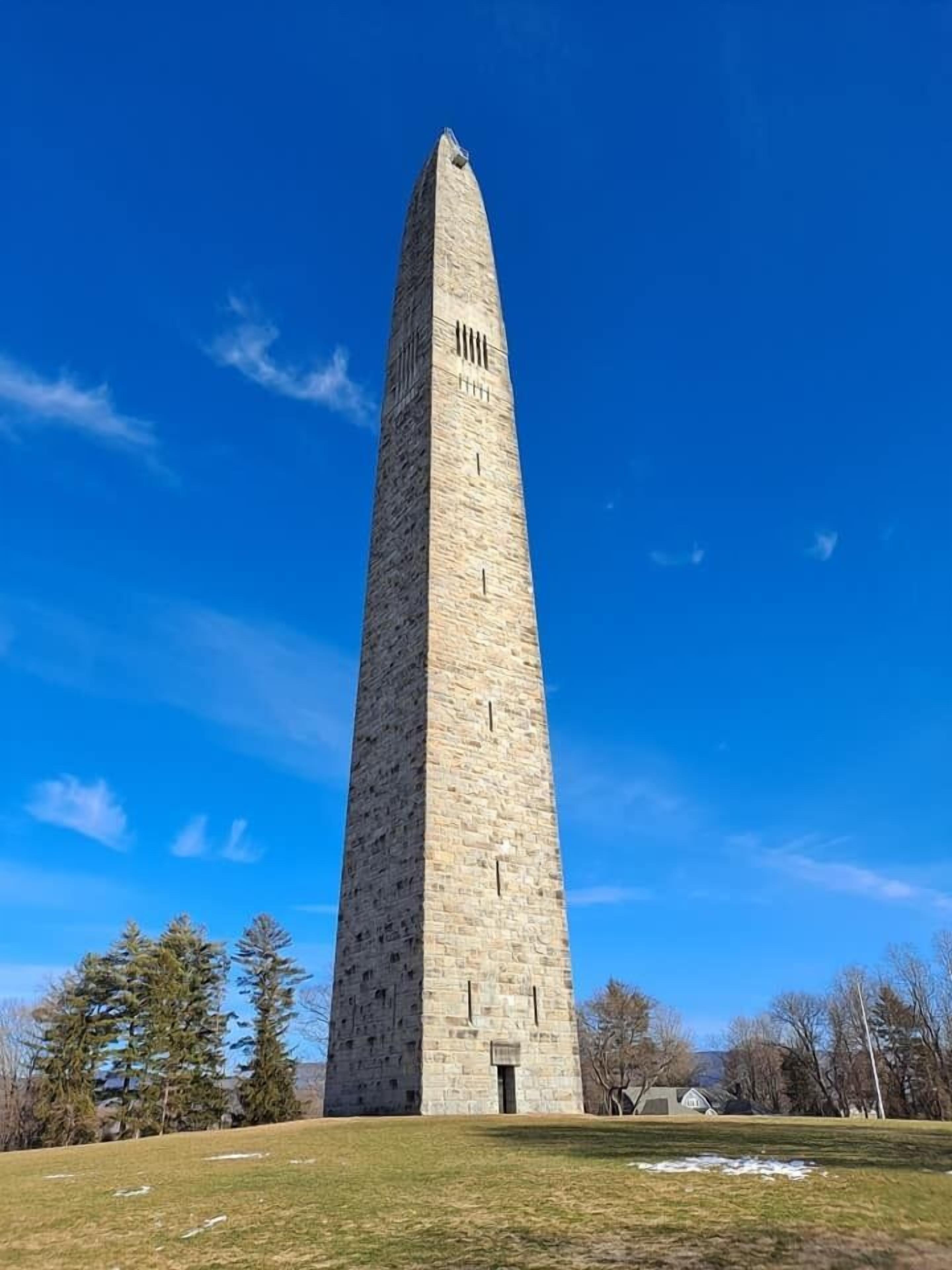 Un monumento alto y esbelto de piedra con una parte superior puntiaguda, rodeado por un campo cubierto de hierba y árboles.