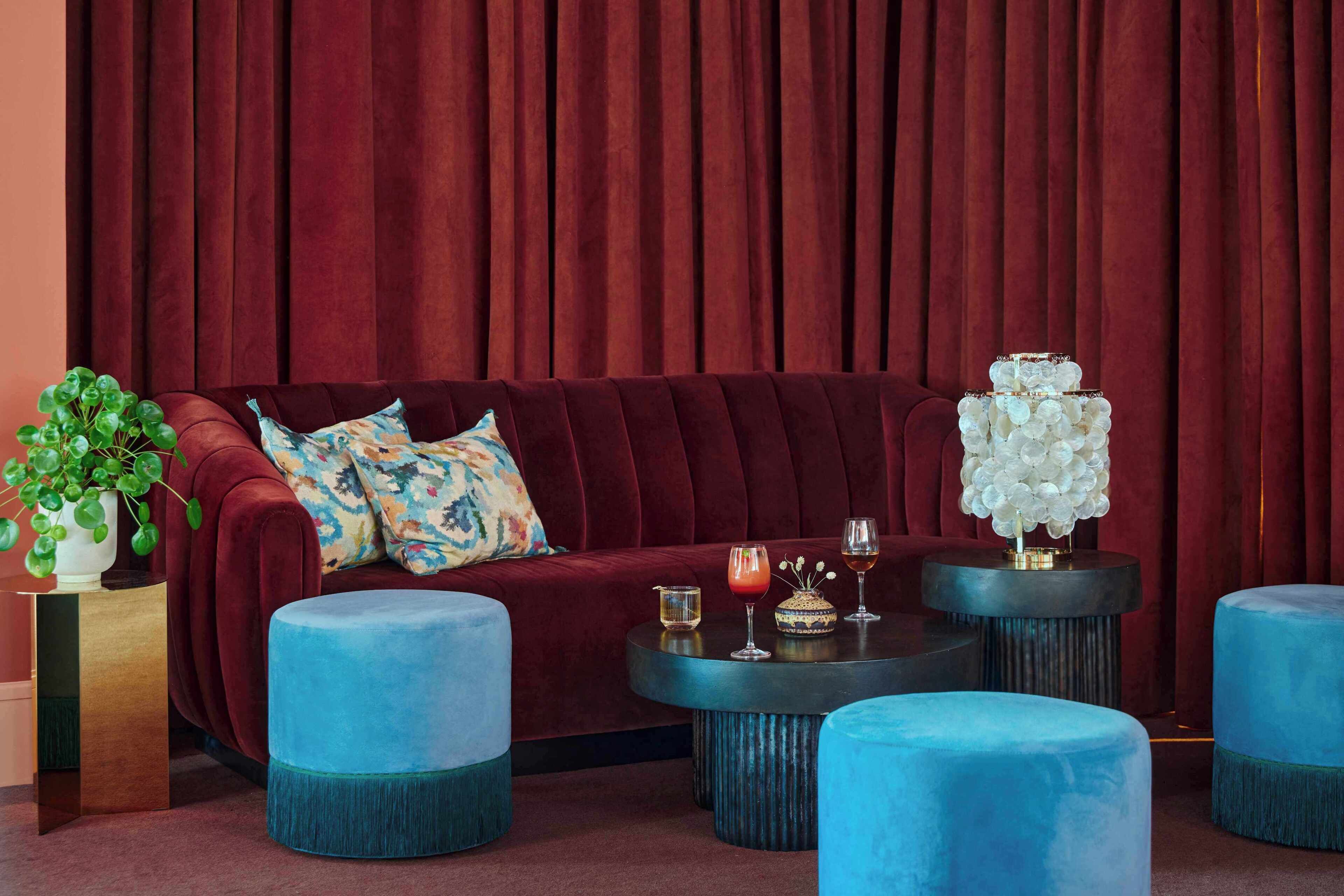 Una habitación con un sofá de terciopelo rojo, otomanas azules y una mesa de centro redonda con bebidas y un florero.
